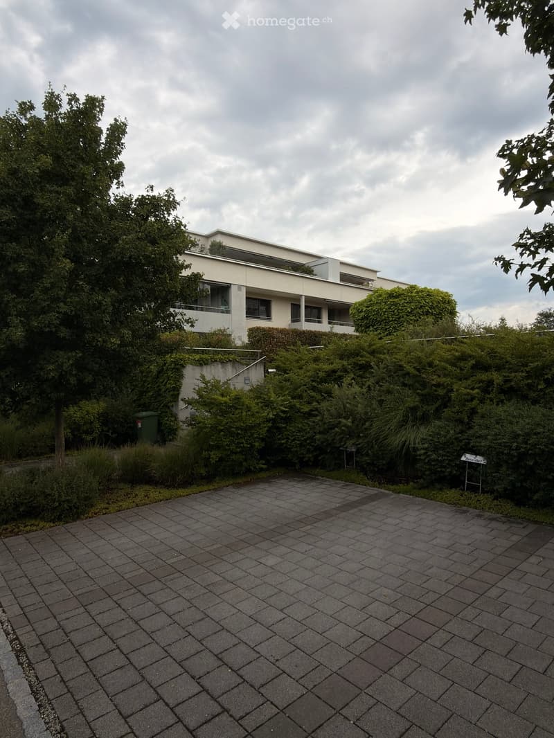 Modern two-story building with white facade, large windows, and raised ground floor surrounded by greenery and a paved driveway.
