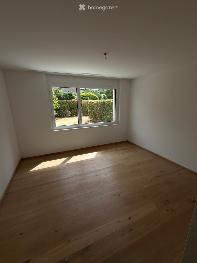 Empty room with light wooden flooring and white walls, featuring a large window with a view of greenery outside.