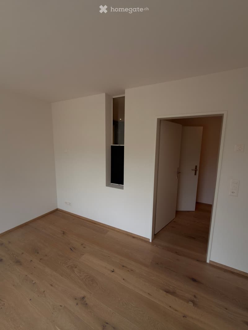 Empty room with light wooden flooring and white walls, featuring a small window and an open doorway leading to another space.