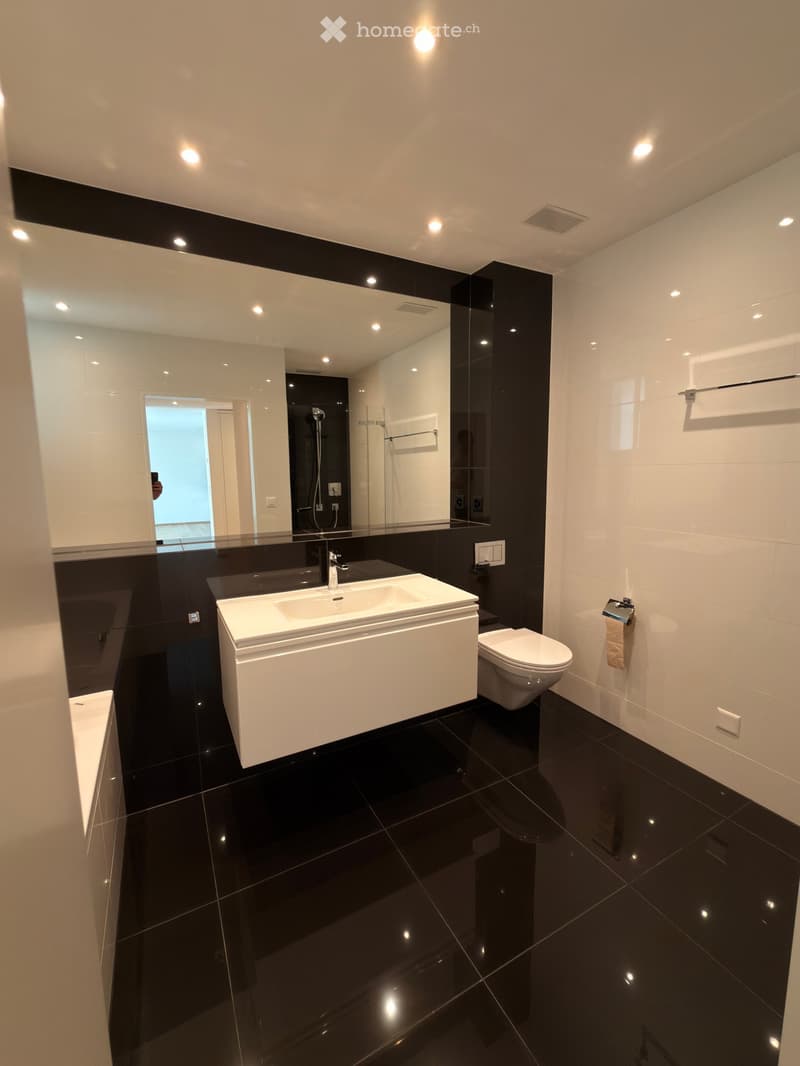 Modern bathroom with black tiled floor and walls, white sink, toilet, large mirror, and modern lighting.