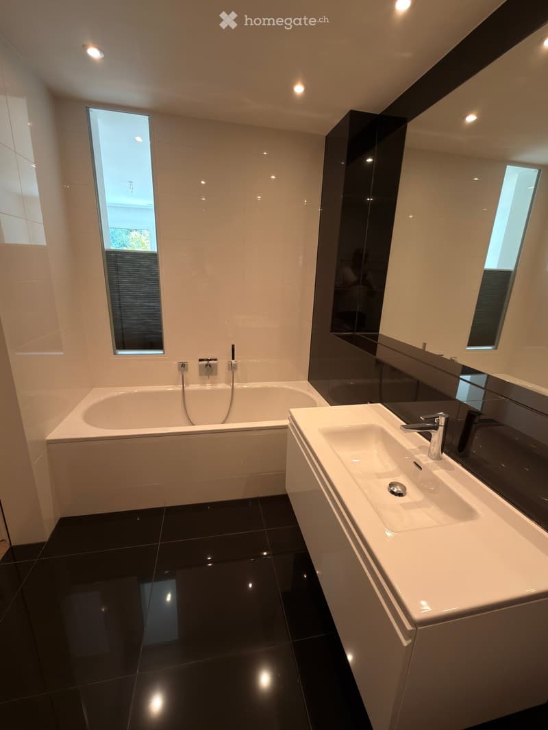 Modern bathroom with a white bathtub, double sink, large mirror, and black tiled floor.