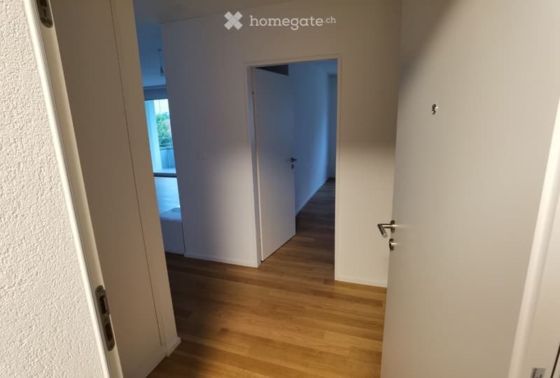 A clean, modern hallway with light wooden flooring and white walls. Doors lead to other rooms, and a glimpse of a room with a window and a bed is visible at the end.