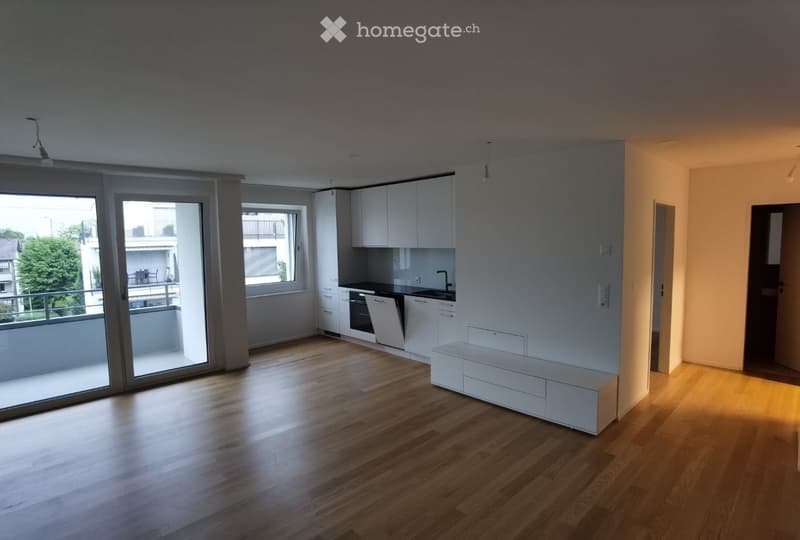 Open-plan living and kitchen area with light wood flooring, white kitchen cabinets, built-in oven and dishwasher, and a white bench. Large sliding glass doors lead to a balcony.