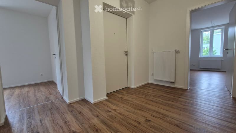 Empty room with wooden flooring, white walls, and a radiator. There are two doors and a window visible.
