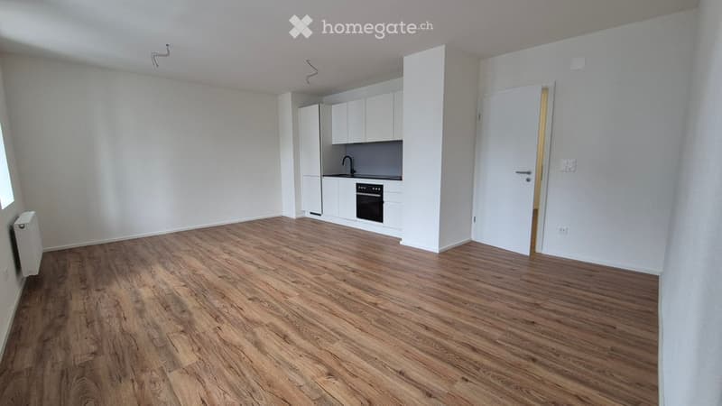 Empty room with wooden flooring, white walls, and a small kitchen area with white cabinets and appliances.