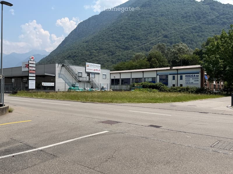 Industrial building complex with multiple signs, surrounded by a grassy area and a road, with a mountain backdrop.