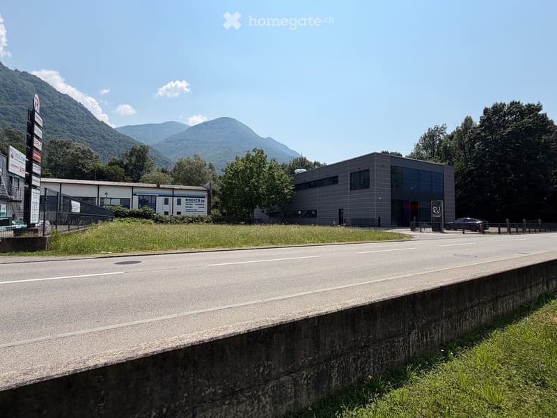 Modern industrial building with glass facade and signage, surrounded by greenery and mountains.