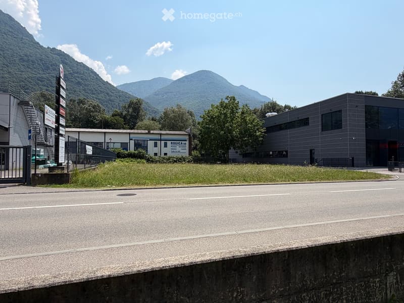Modern industrial building with a flat roof and large windows, surrounded by greenery and mountains.