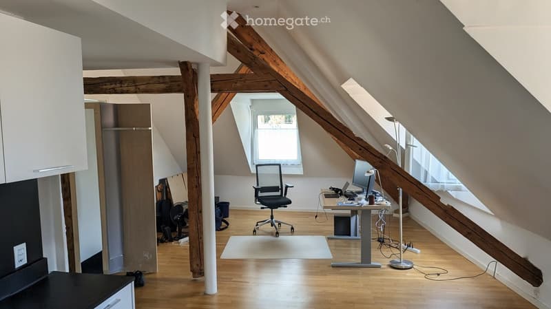 Spacious attic room with wooden beams, large windows, modern furniture including a desk, chair, laptop, and computer monitor.