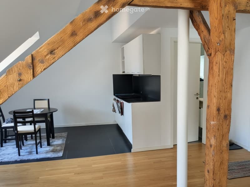 A modern, open-plan living and dining area with a black dining table and chairs, a sleek kitchen counter with cabinets, and exposed wooden beams on the ceiling.