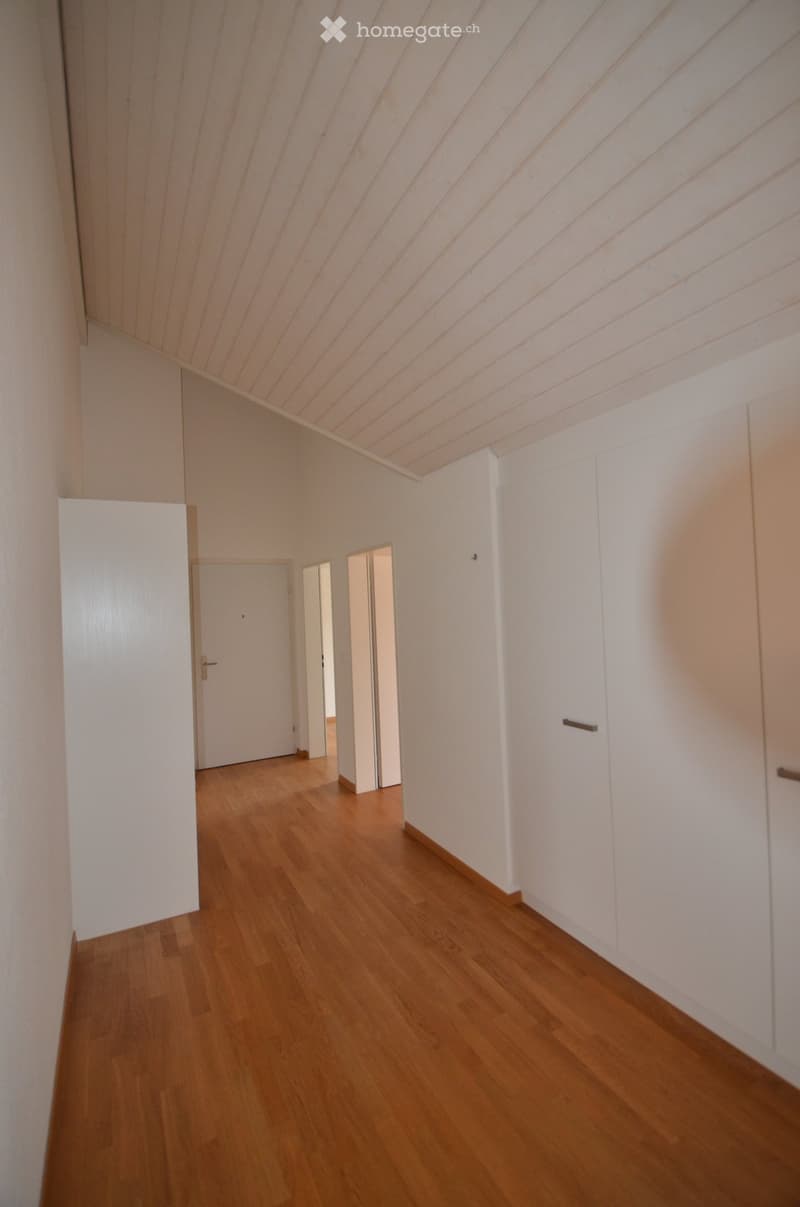 Empty hallway with wooden flooring, white walls, and white cabinetry on one side. The ceiling has a slanted design with wooden paneling.