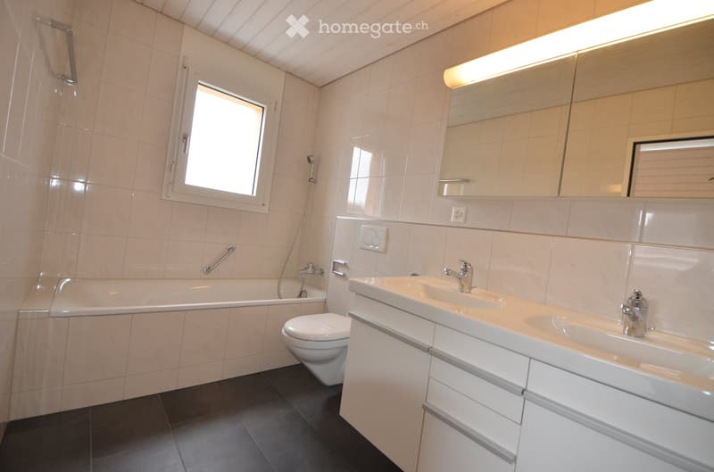 Modern bathroom with a bathtub, toilet, double sink, and large mirror. Features tiled walls and a window for natural light.