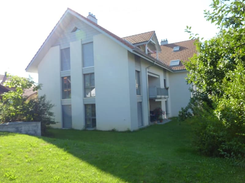 Two-story house with light beige walls, large windows, and a balcony on the upper level. The roof is covered with red tiles, and the house is surrounded by a well-maintained lawn and trees.