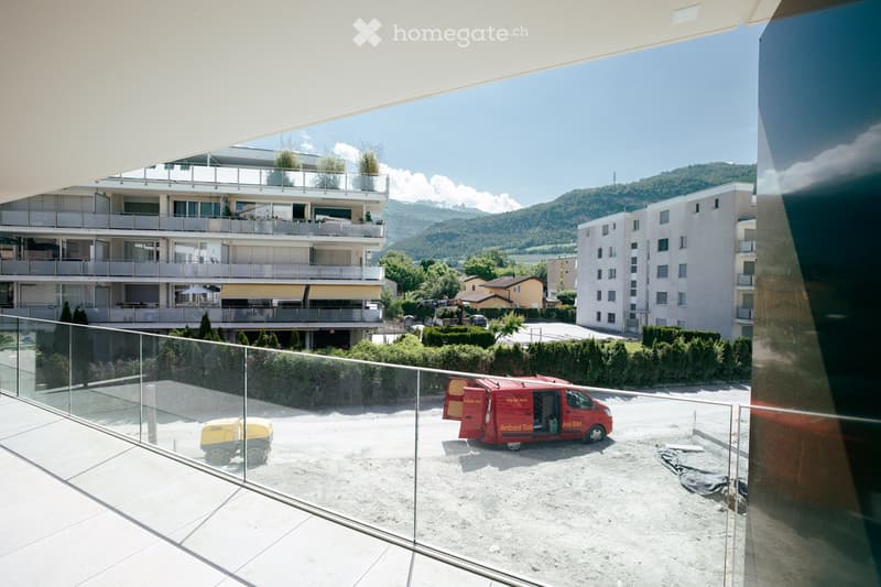 Balcony overlooking a construction site with a red van, yellow machinery, and surrounding buildings with balconies and greenery.