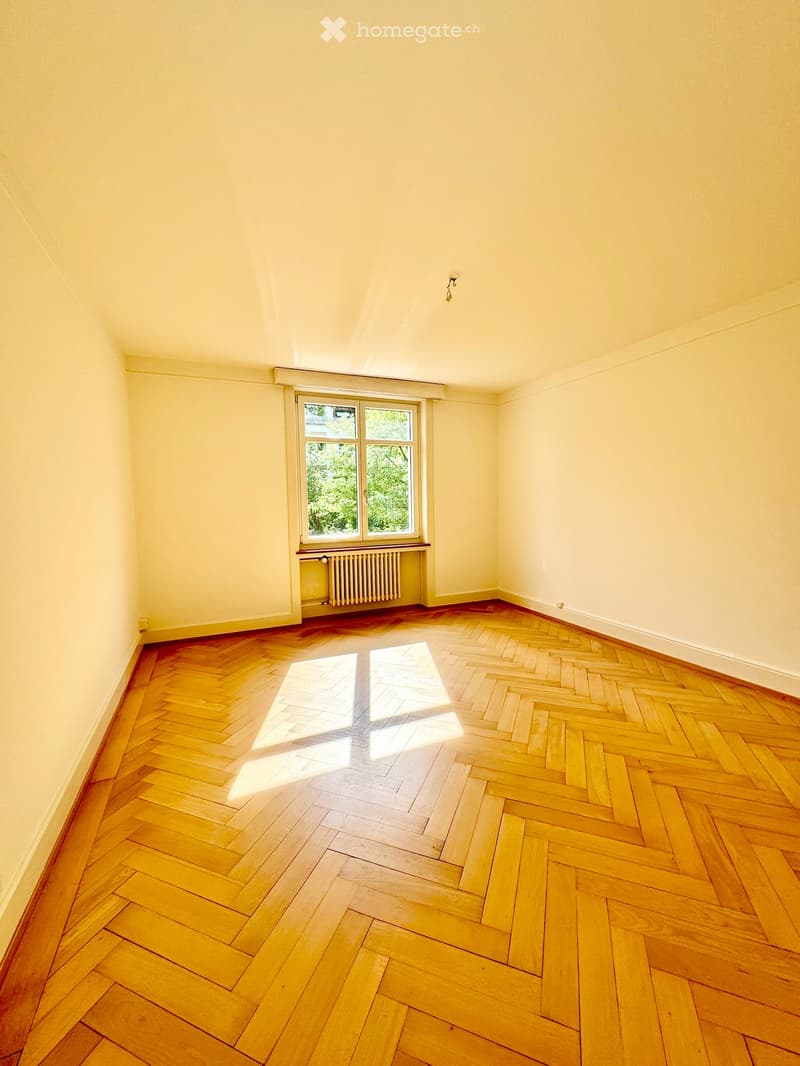 Empty room with wooden herringbone flooring, light yellow walls, and a single window with a radiator below it.