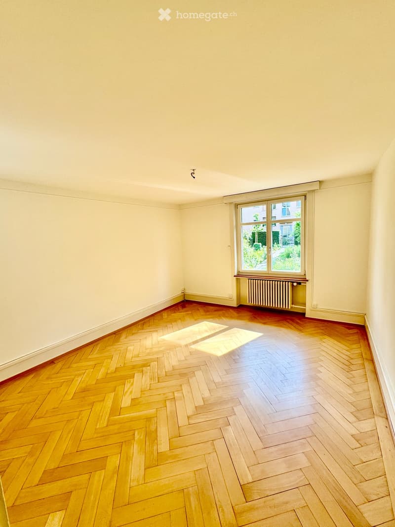 Empty room with wooden herringbone flooring, white walls, and a large window with a radiator below it.