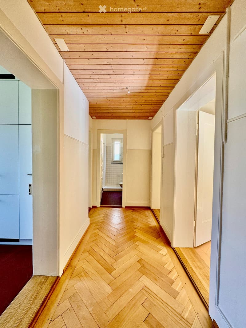 A long, narrow hallway with herringbone-patterned wooden flooring, light-colored walls, and a wooden ceiling. Doors line both sides, leading to various rooms.