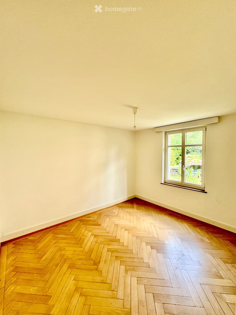 Empty room with light-colored walls, wooden herringbone flooring, and a large window with a view of greenery outside.
