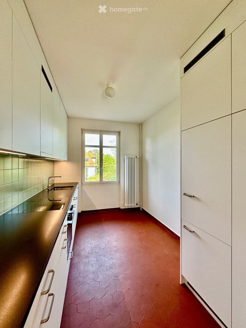Empty kitchen with white cabinets, dark countertops, and a red hexagonal tile floor. A window with a view of greenery is present, along with a radiator and a ceiling light fixture.