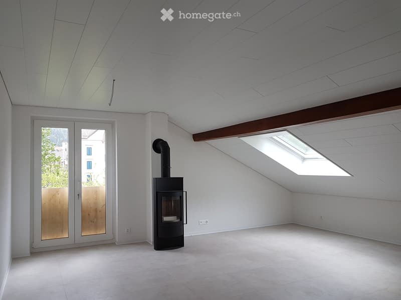 Empty attic space with white walls, a skylight window, and a wood-burning stove with a chimney. The room has a high, slanted ceiling and a large sliding glass door leading outside.