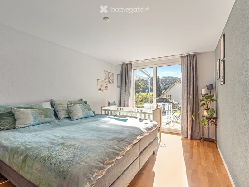 Bright bedroom with a large bed, nightstand, lamp, and a plant near the window. The room features wooden flooring and large windows with curtains, offering a view of the outdoor area.