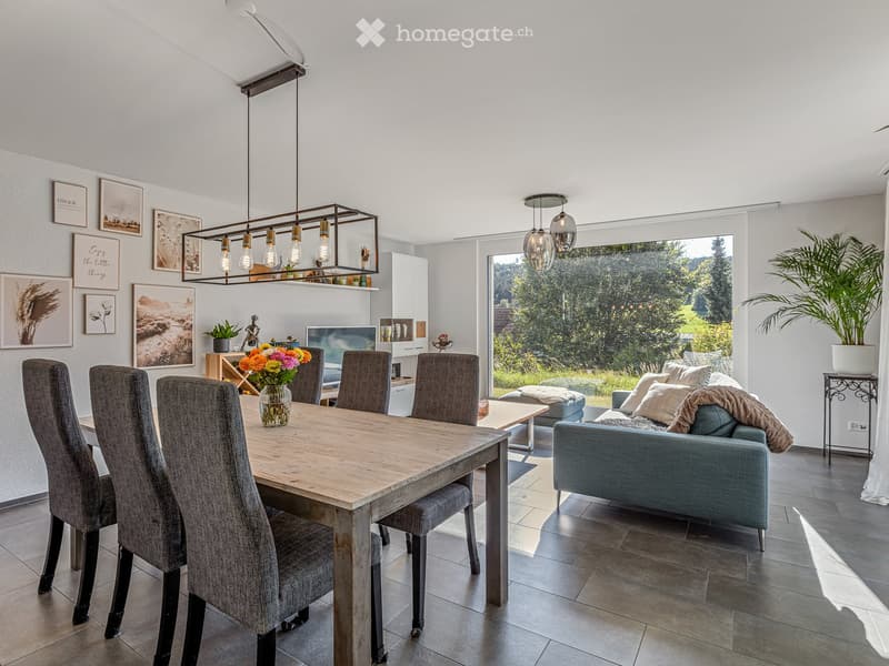 Modern living and dining area with a grey sofa, wooden dining table, six chairs, a vase with flowers, a television, and a potted plant. Large windows provide natural light, and the space is decorated with picture frames and a hanging light fixture.
