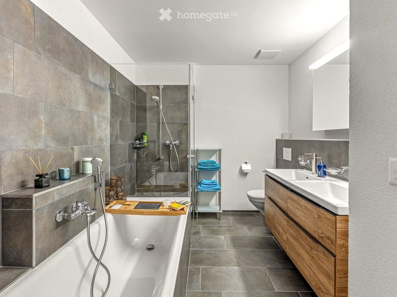 Modern bathroom with a bathtub, shower, double sink, and wooden cabinet. Includes a shelf with towels and decorative items.