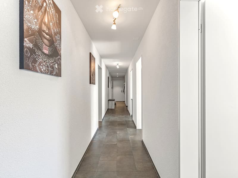 Clean, modern hallway with white walls, tiled floor, and recessed lighting. Features several doors and framed artwork on the walls.