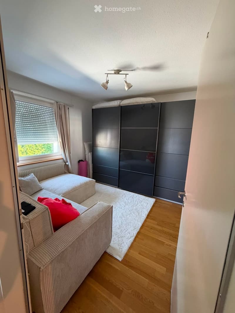 Cozy living room with a sofa, ottoman, large wardrobe, and a window with curtains. Wooden flooring and a white rug.