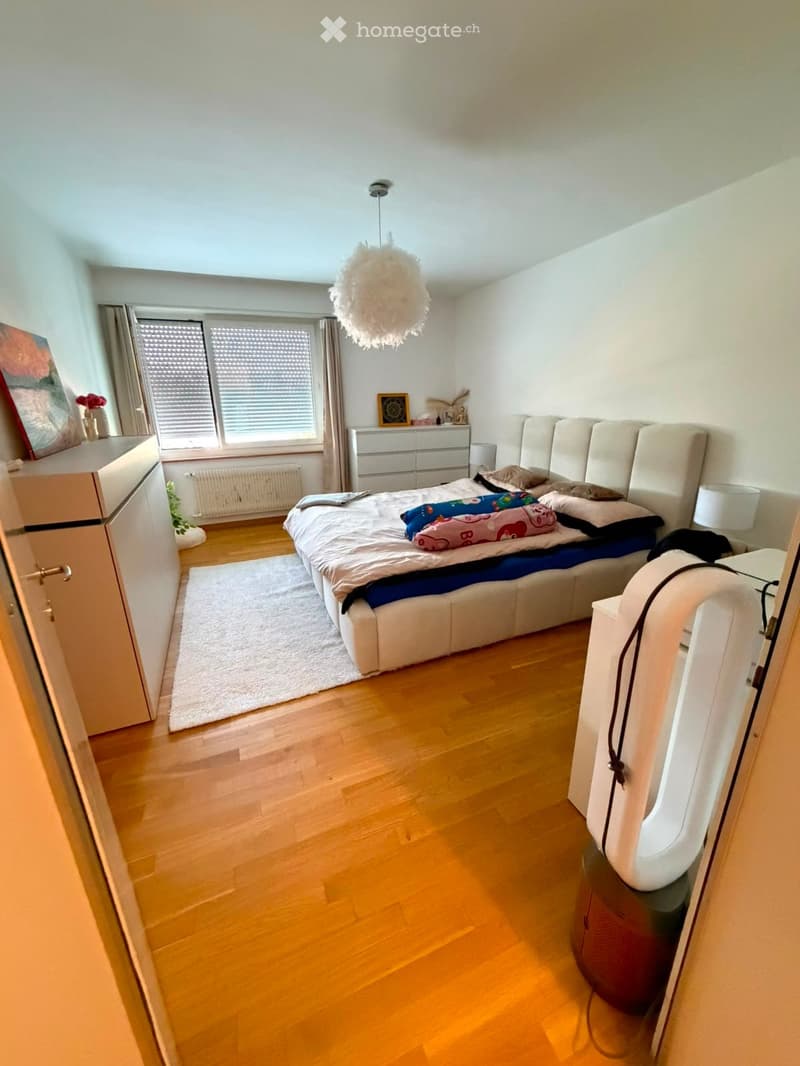 Modern bedroom with wooden flooring, large bed with white headboard, nightstand with lamp, dresser, and a hanging decorative light fixture.