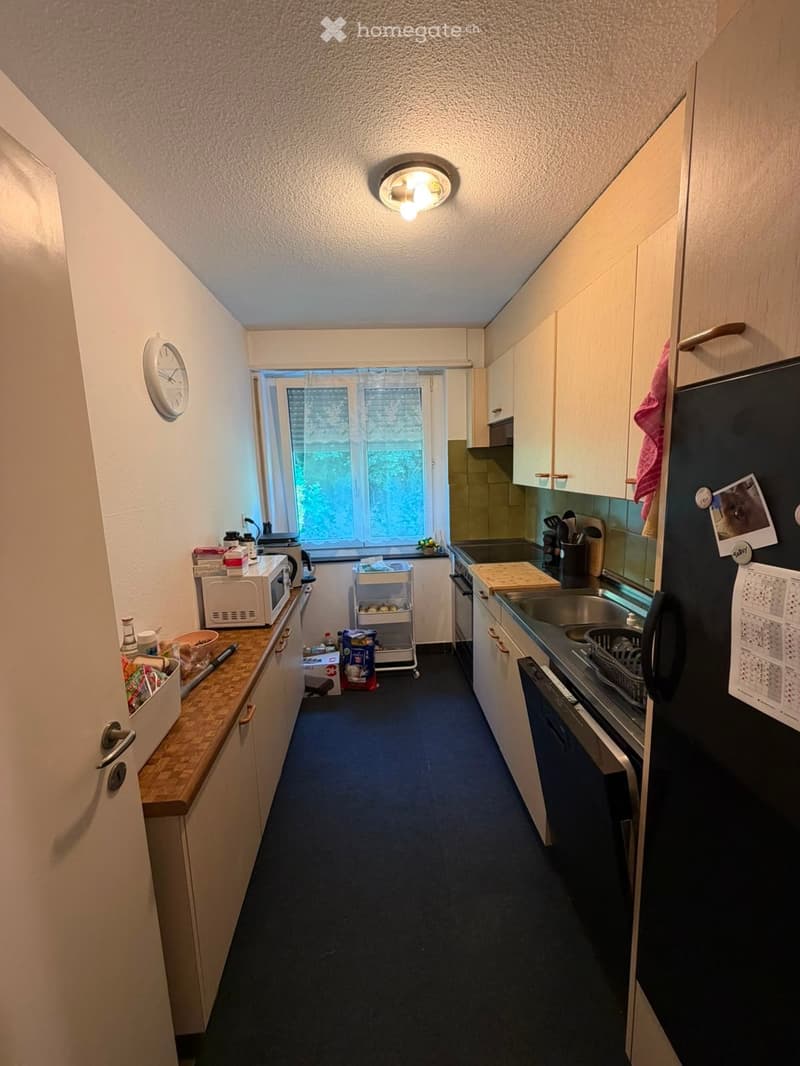 A compact kitchen with white cabinets, a dark refrigerator, microwave, oven, sink, and countertop. A small window provides natural light, and a clock is mounted on the wall.