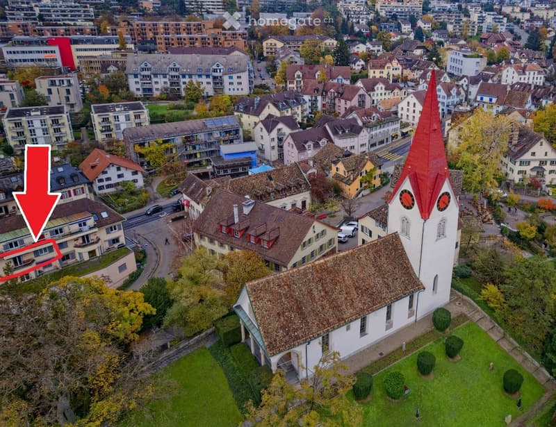 Church with red roof, surrounded by houses, trees, and grass, located in a city