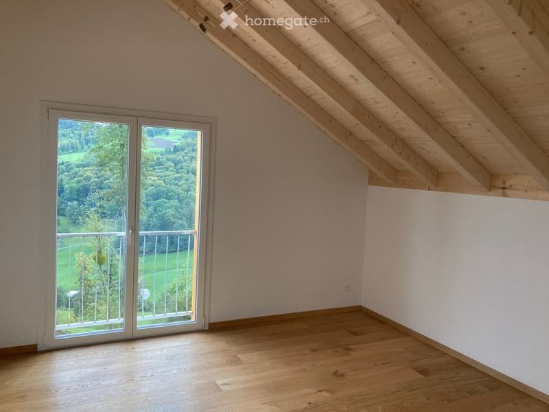 Empty attic room with wooden slanted ceiling, wooden floor, large sliding glass door leading to a balcony with scenic view of green landscape.