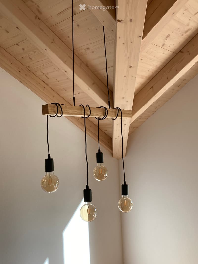 An unfurnished room with exposed wooden beams and a modern pendant light fixture featuring four vintage-style bulbs.