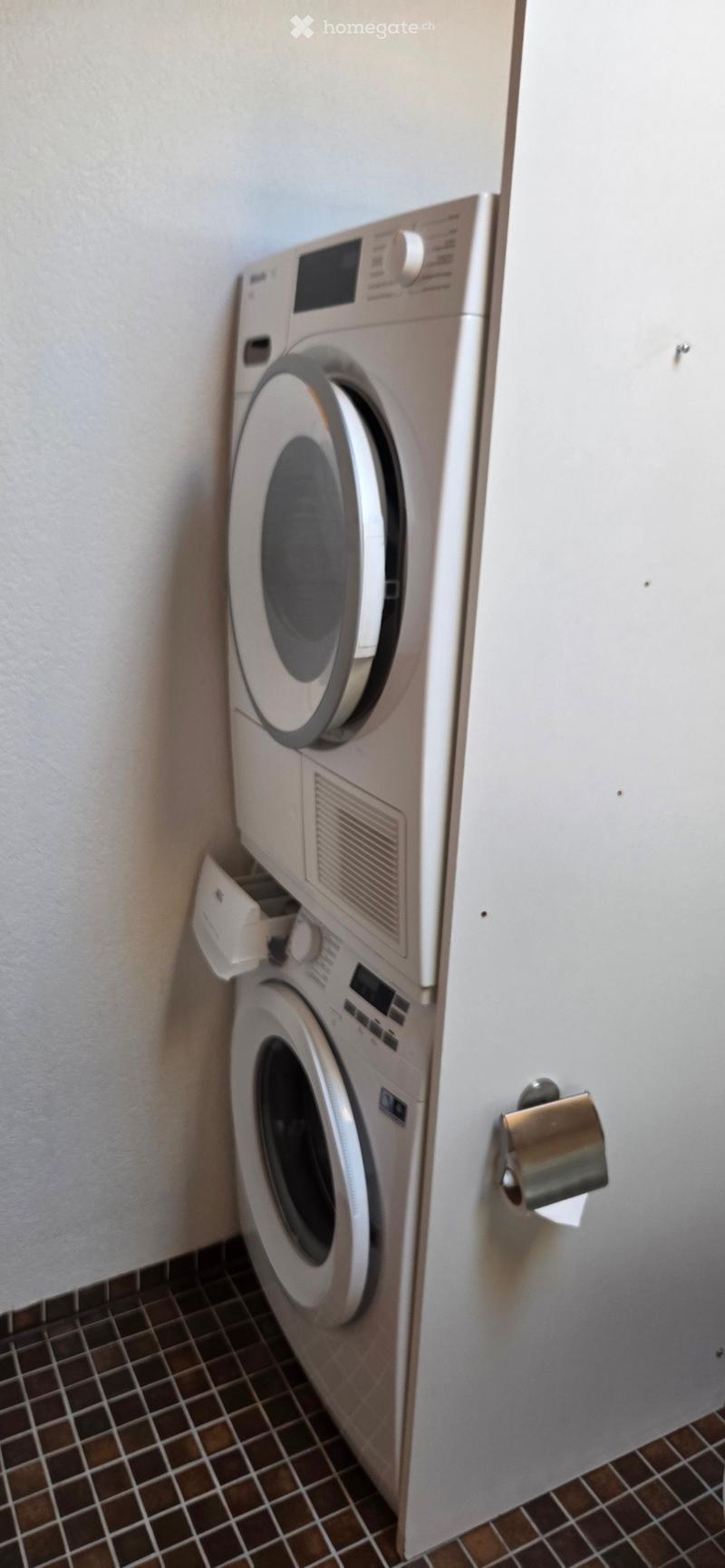 A compact bathroom with a stacked washing machine and dryer unit, tiled floor, and a toilet paper holder on the wall.