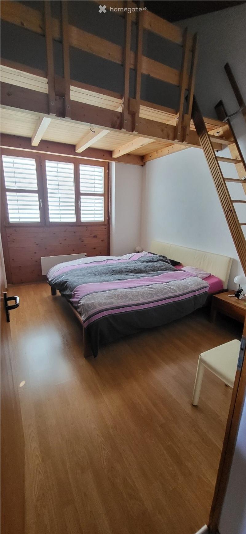Cozy bedroom with wooden flooring, a double bed with a grey and pink duvet, a nightstand with a lamp, and a small white stool near the window.