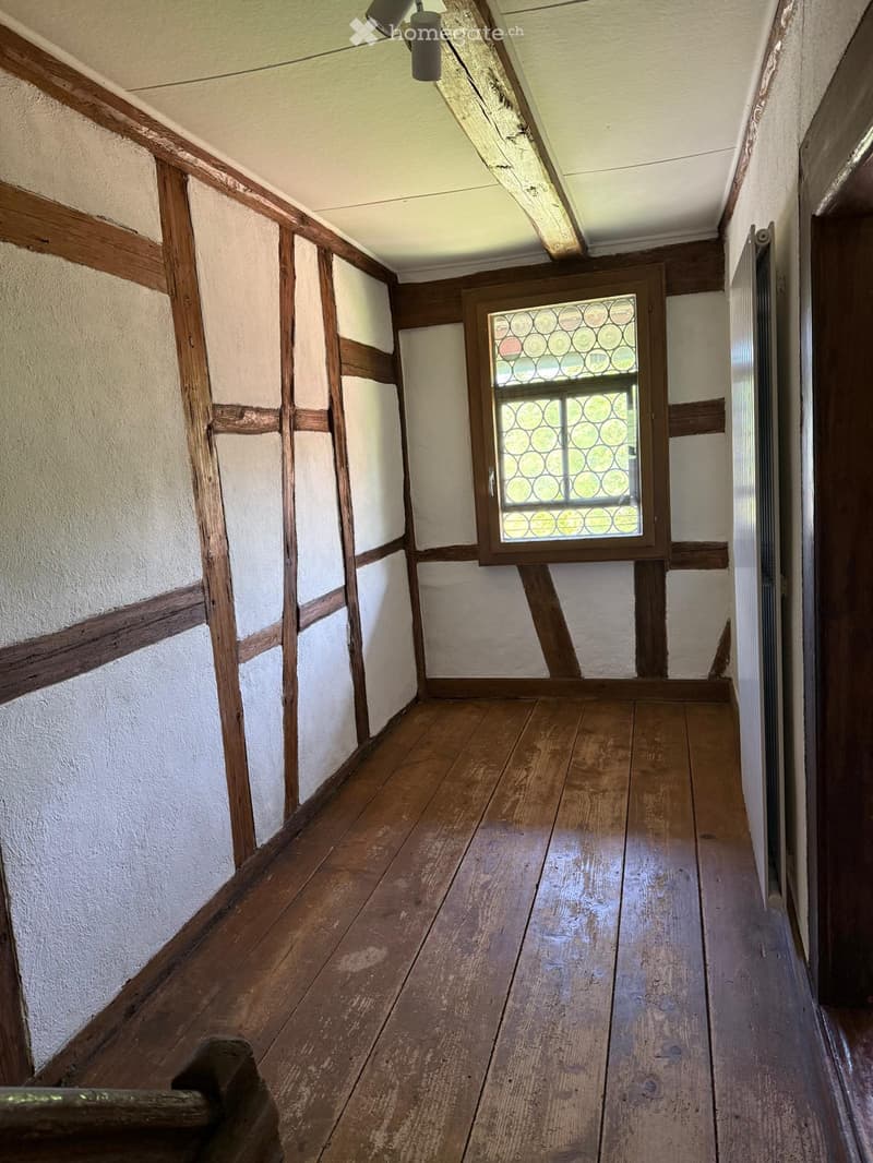 Empty wooden-floored corridor with exposed wooden beams and white plaster walls, featuring a single stained-glass window at the far end.