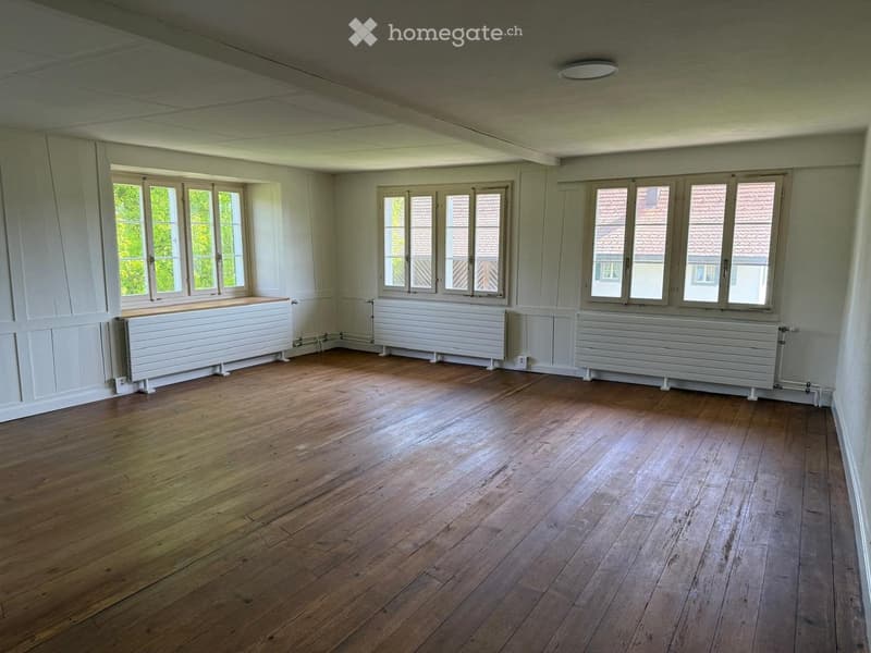 Empty room with wooden flooring, white walls, large windows, and radiators along the walls.