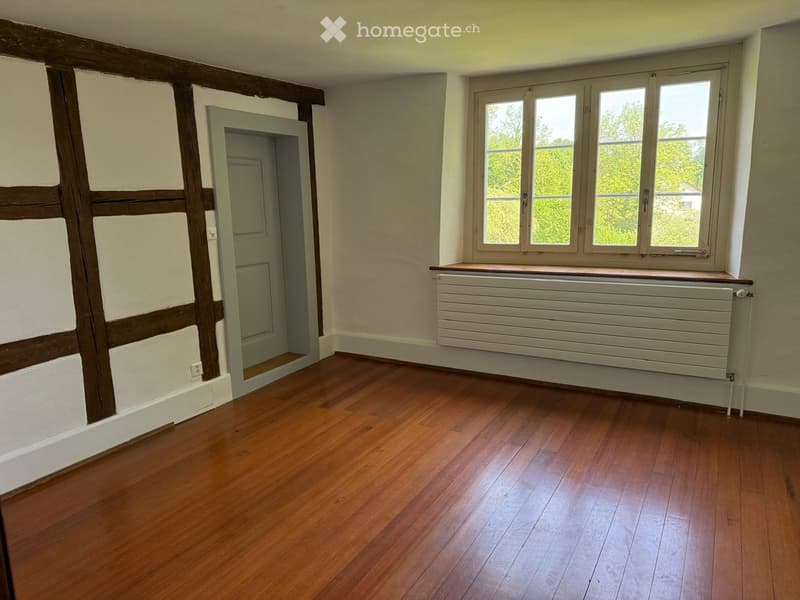 Empty room with wooden flooring, exposed wooden beams on one wall, a radiator, and a door leading to another space.