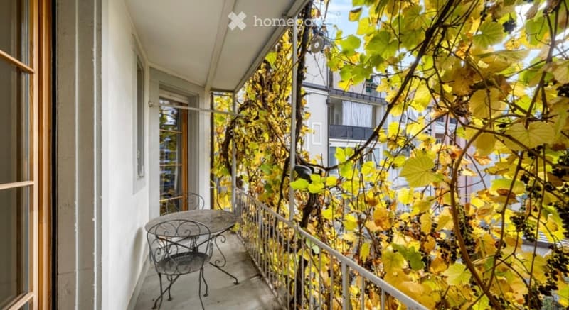 Balcony with a metal table and chair, covered with grapevines, overlooking a cityscape.
