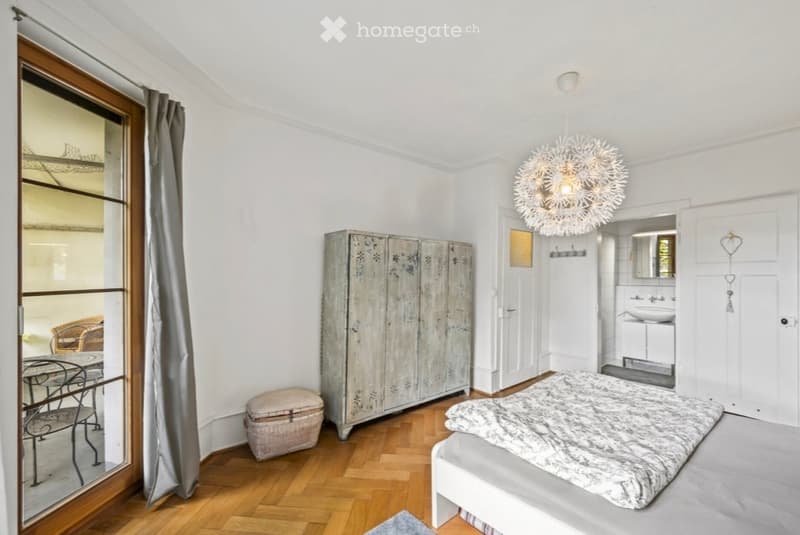 Bright bedroom with wooden flooring, a large bed, a vintage wardrobe, a small table, and a chandelier. A small bathroom is visible through an open doorway.