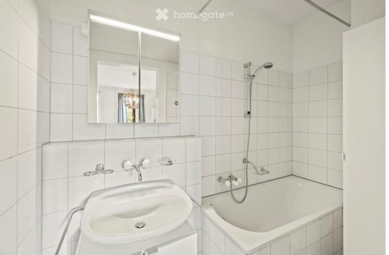 Modern bathroom with white tiled walls and floor, featuring a sink, bathtub, and a large mirror above the sink.
