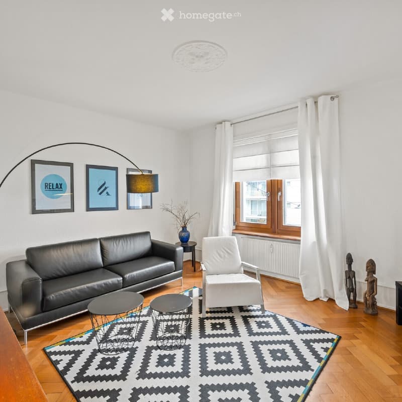 Modern living room with a black leather sofa, white armchair, two small round tables, a patterned rug, and a floor lamp. The room features large windows with white curtains and several picture frames on the wall.