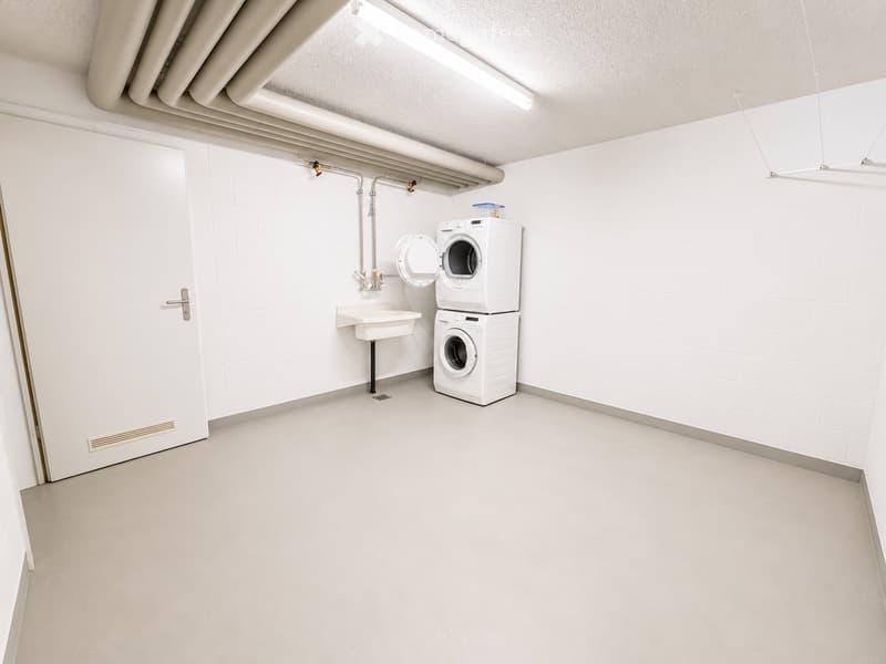 A clean, modern laundry room with white tiled walls, a washing machine, a dishwasher, and a sink.