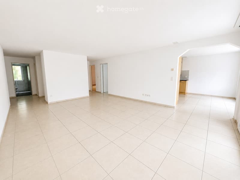 Empty room with tiled floor and white walls, featuring a doorway leading to another room and a small kitchen area visible in the background.