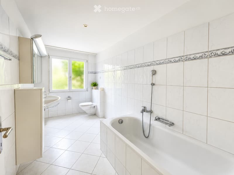 Bright bathroom with white tiles, a bathtub, toilet, sink, and a small plant on the windowsill. The room features a large window with frosted glass and a modern showerhead.