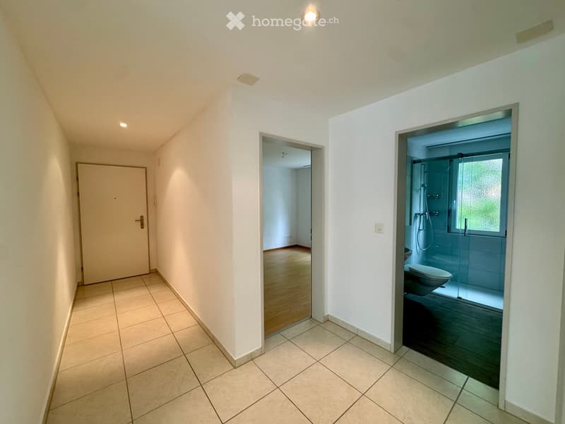 Empty hallway with white walls, tiled floor, two doors, one leading to a bathroom with a shower and toilet, the other leading to an empty room with wooden floor.