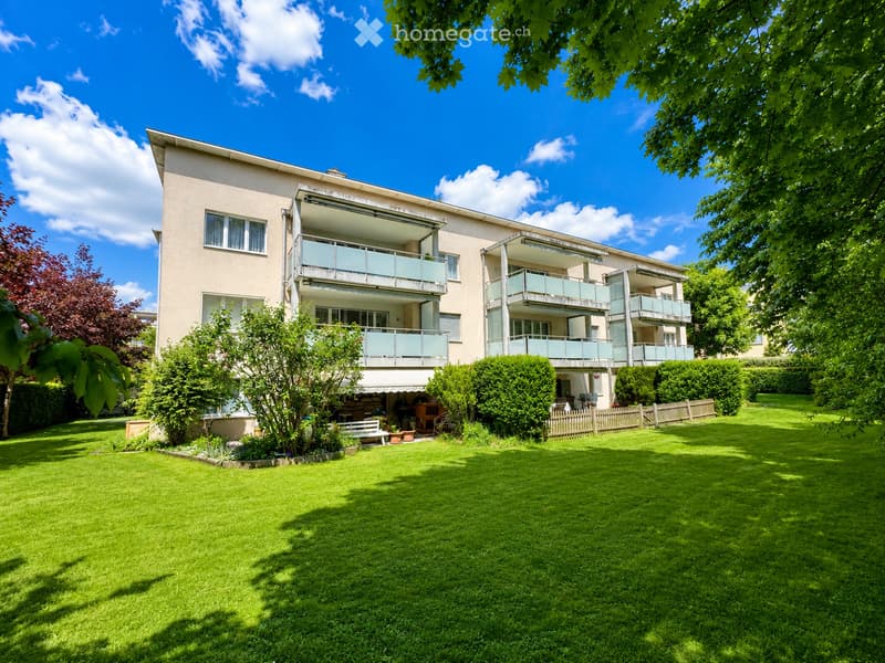 A modern multi-story residential building with balconies, surrounded by a well-maintained lawn and trees.