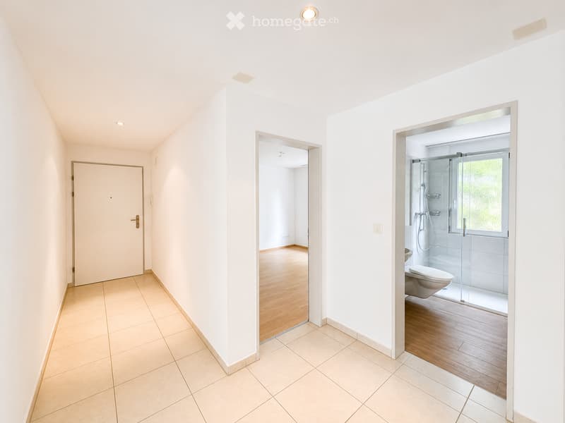 Bright hallway with tiled floor, white walls, and recessed lighting. Opens to a room with wooden flooring and a bathroom featuring a glass-enclosed shower and toilet.