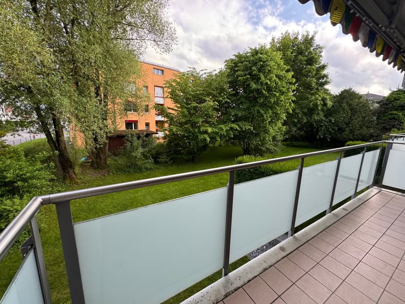Balcony with glass railings, tiled floor, overlooking a grassy area with trees